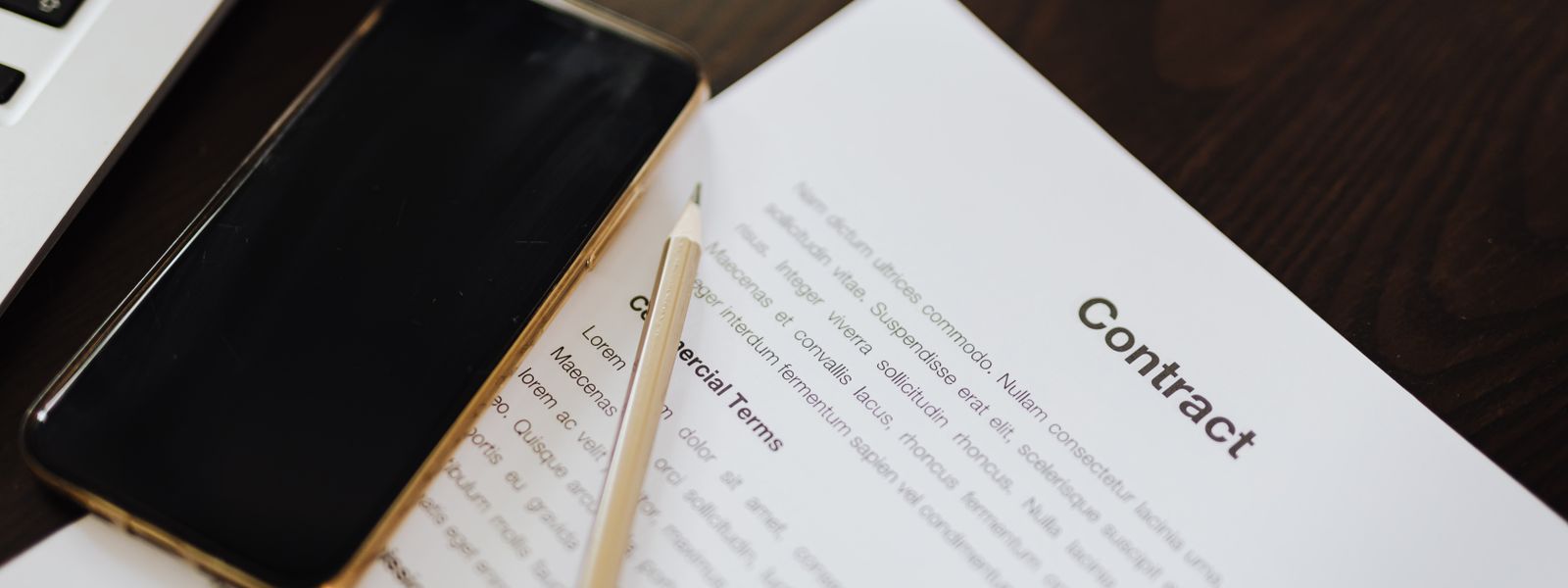 Laptop, smartphone and a printed contract document with a pen lying on a wooden office desk