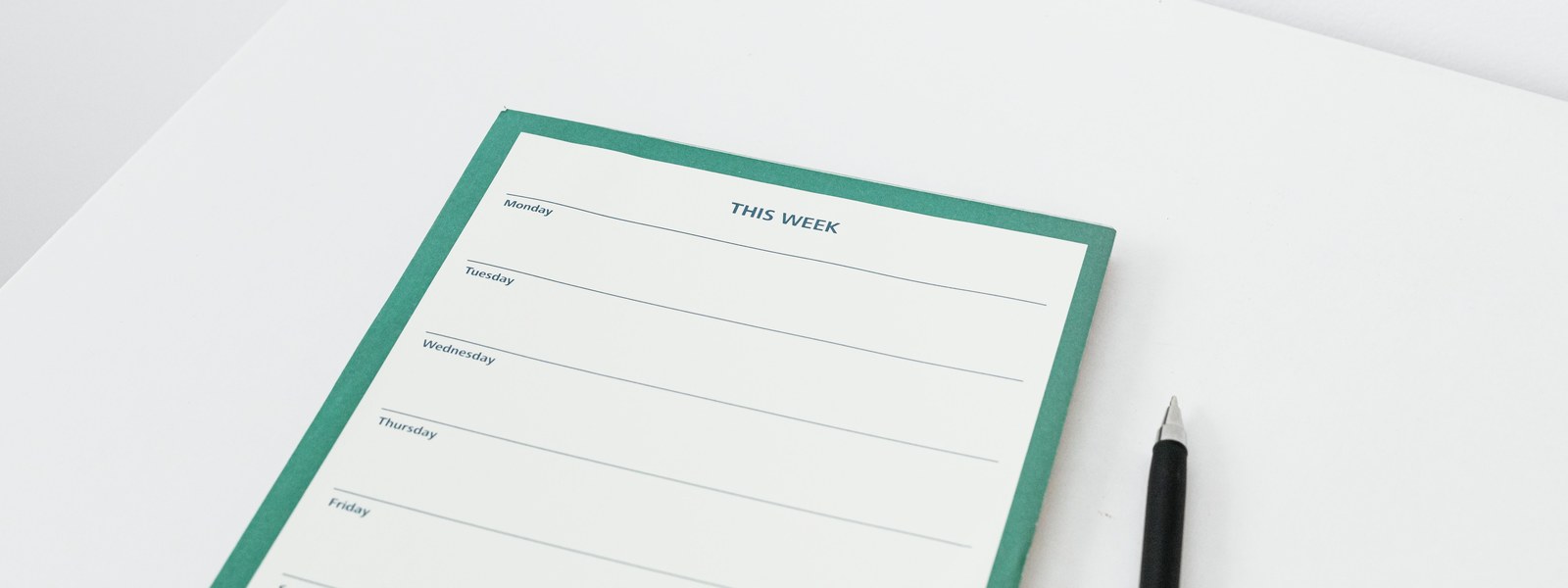A blank green-bordered weekly planner headed This Week, with a ruled line for each day, on a white desk beside a black pen