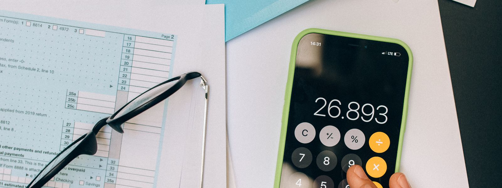 Hand using a phone calculator on a desk beside paperwork and a pair of glasses