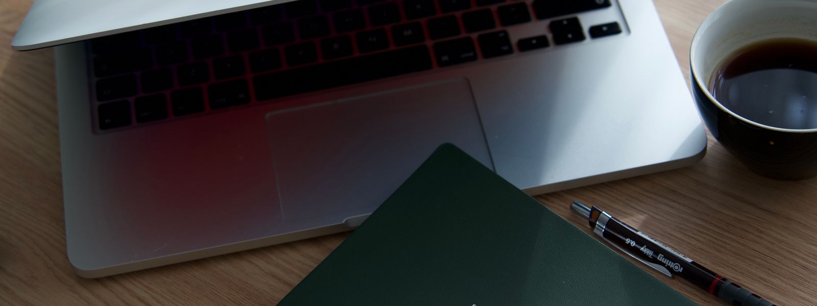Open laptop, a dark green notebook, a fine-liner pen and a cup of black coffee on a wooden desk