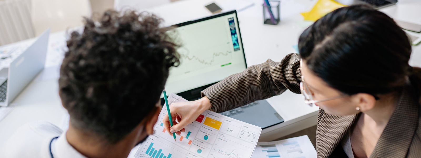 Two consultants in an office reviewing printed charts and a laptop across a desk during a client meeting