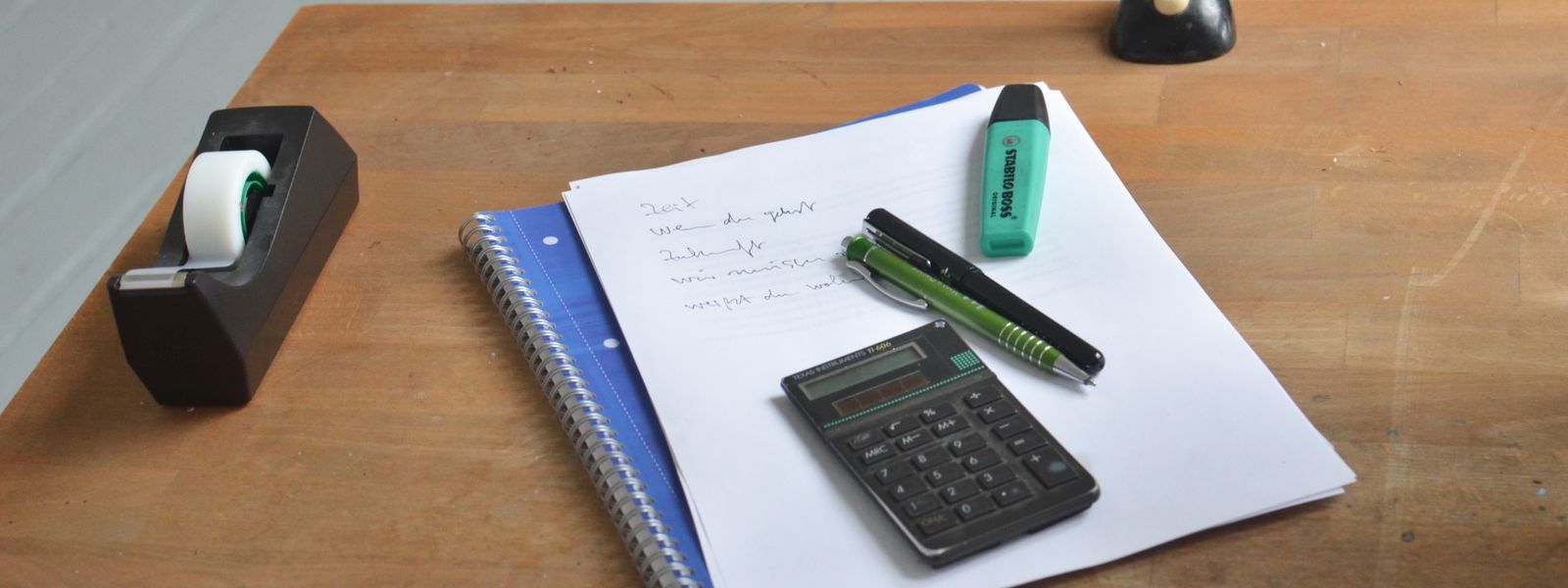 Overhead view of a wooden office desk with printed working papers, a small calculator, pens and a highlighter laid out ready for a review