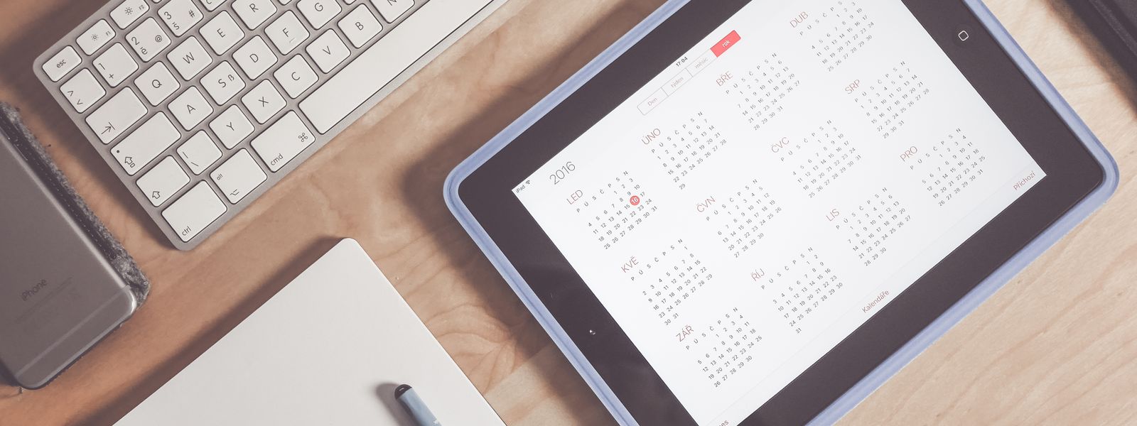 Tablet on a wooden desk showing a year of calendar months beside a keyboard, phone and notebook