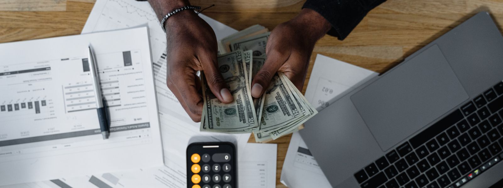 A person counting US dollar bills at a wooden desk beside a calculator, a laptop and financial paperwork