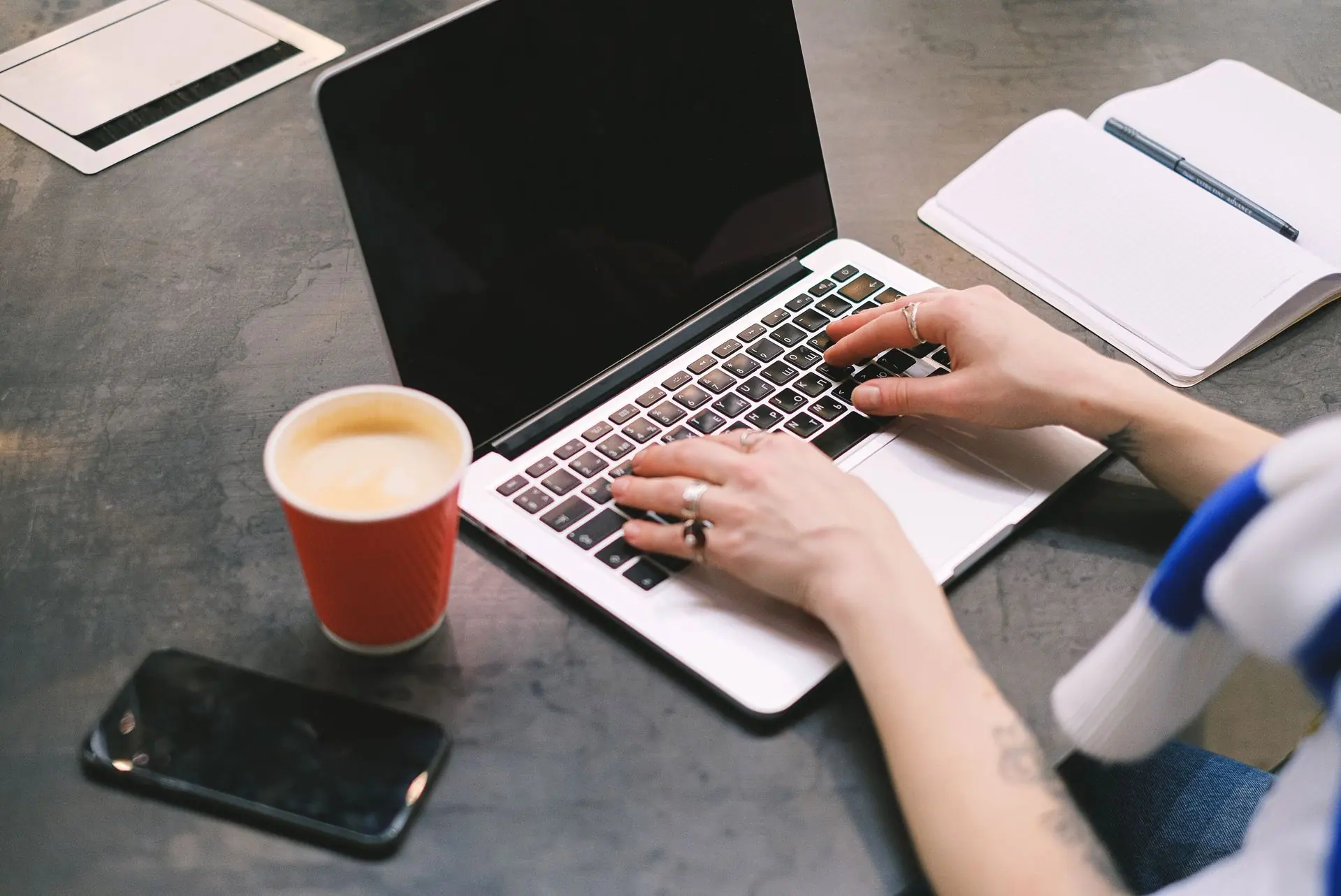 A person typing on a laptop at a desk, with a takeaway coffee, a phone and an open notebook beside them