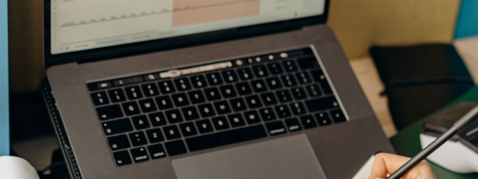 A laptop showing a spreadsheet on a desk, beside a hand making notes with a pen
