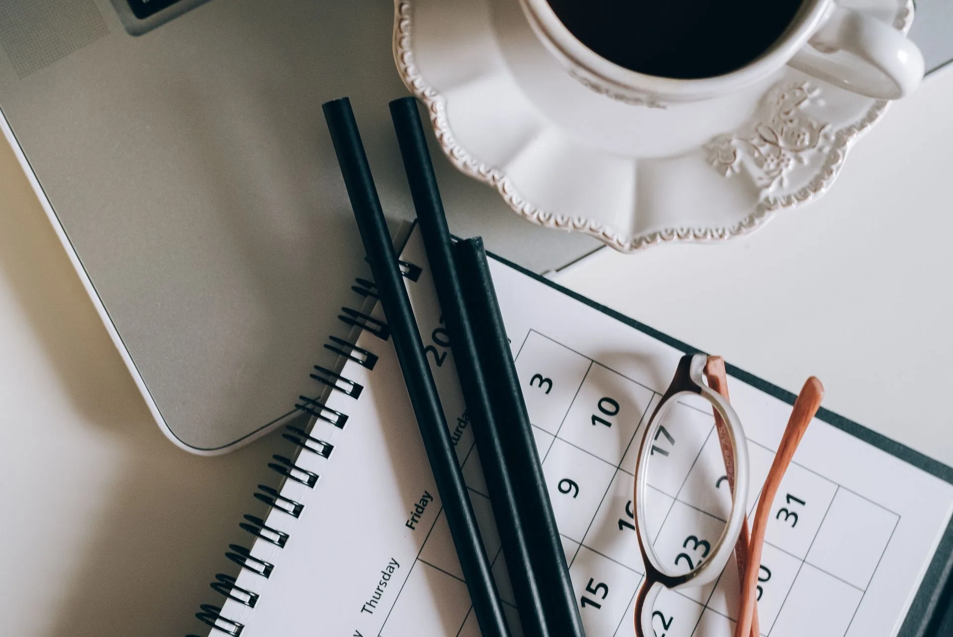 A monthly calendar with a pair of glasses and pencils resting on it, next to a laptop and a cup of coffee